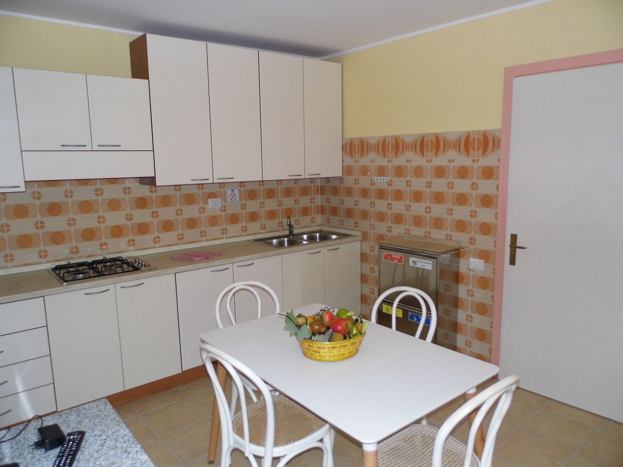 Una pequeña cocina con armarios blancos, estufa a gas, un lavamanos y una mesa de comedor con sillas. Hay una cesta de frutas sobre la mesa.