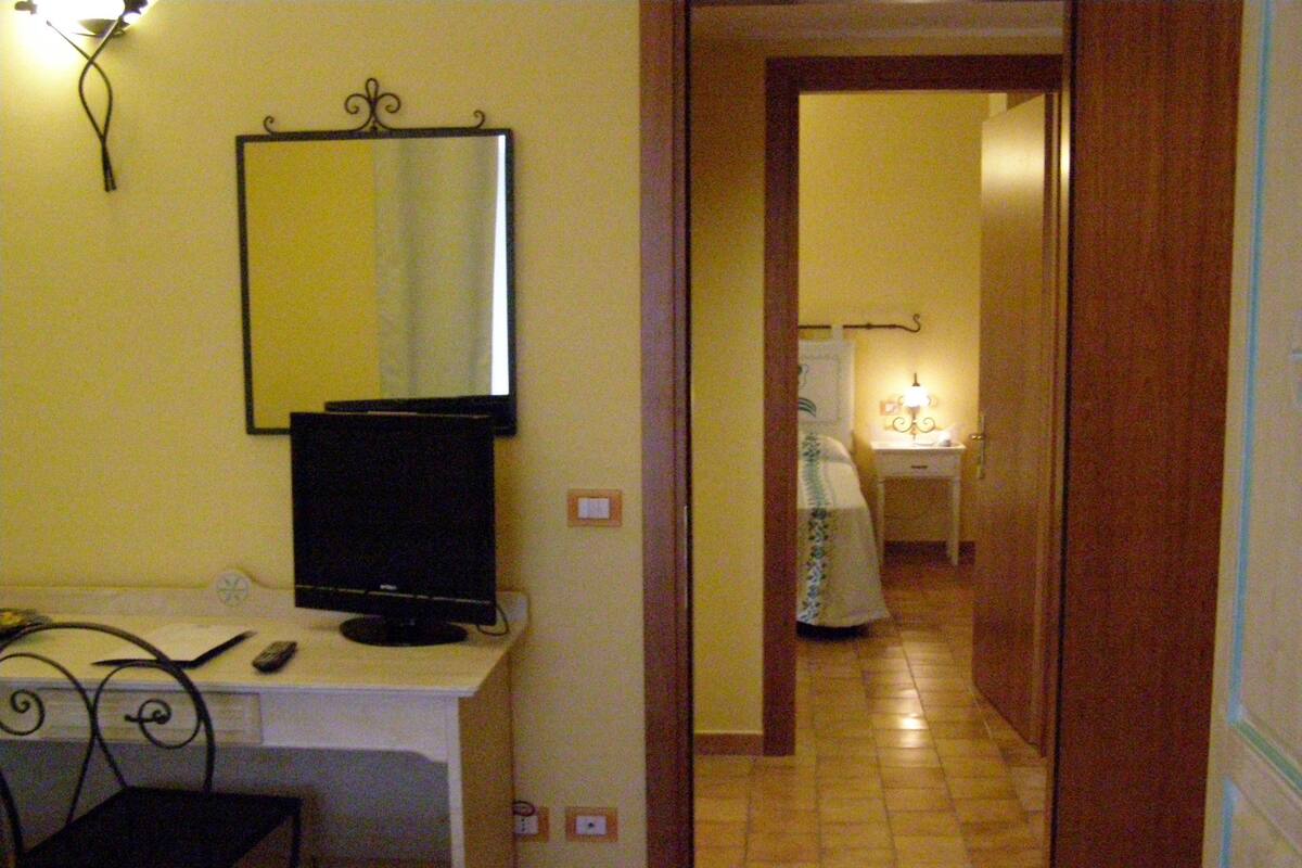 Habitación de hotel con cama, escritorio con computadora, televisor y espejo. Hay una puerta que da a otra habitación.