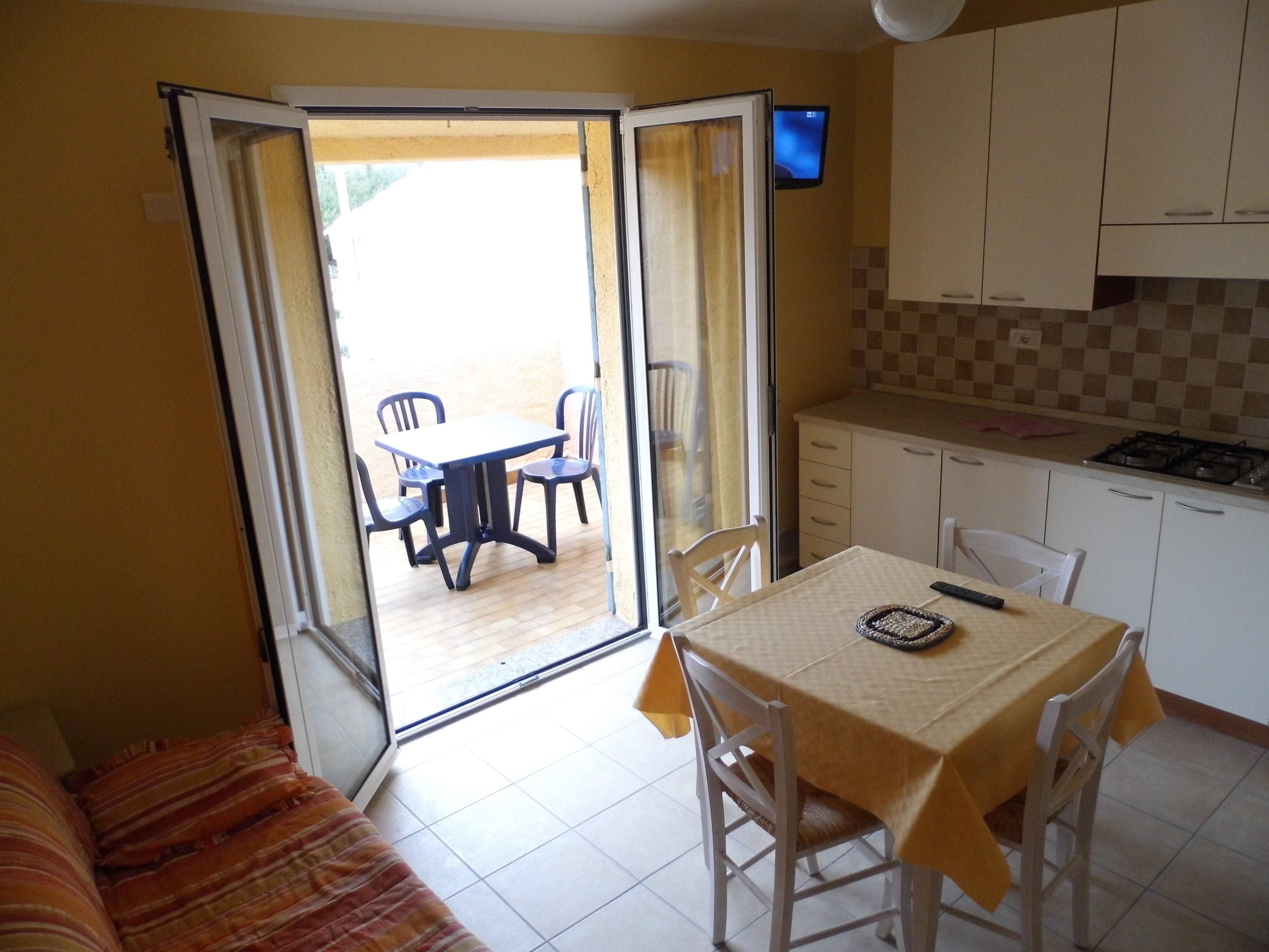 A compact kitchen with white cabinets and a tiled backsplash, a dining area with a table and chairs, and a sliding glass door leading to an outdoor patio with a table and chairs.