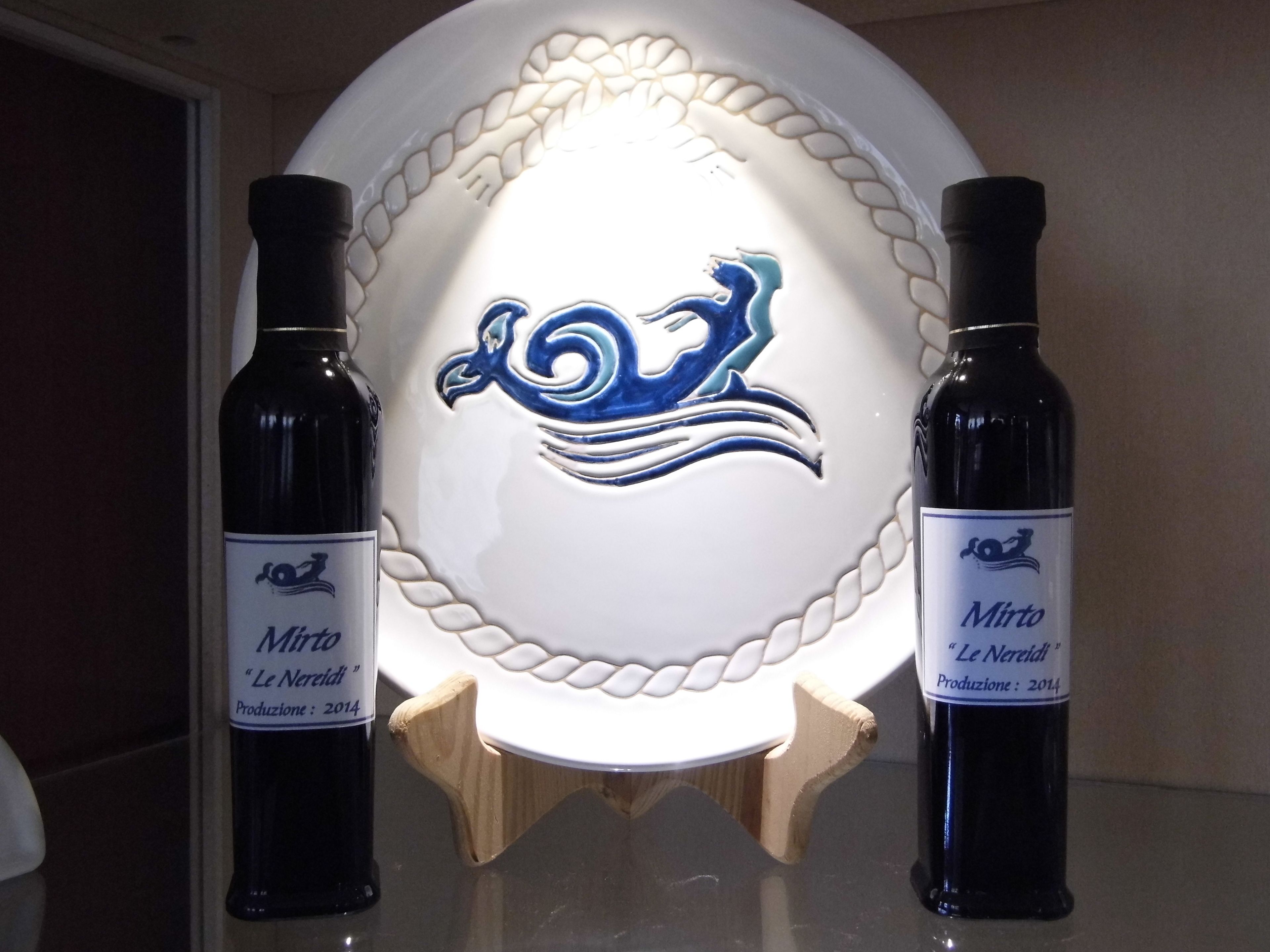 Dos botellas de vino Mirto "Le Neretti" sobre una superficie reflectante, con un plato decorativo de diseño azul al fondo.