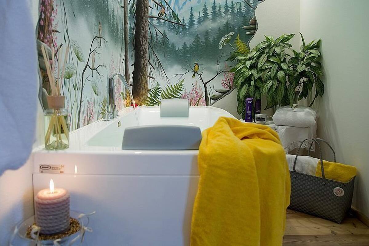 Un baño con bañera, un mural de un bosque, una vela y una toalla amarilla.