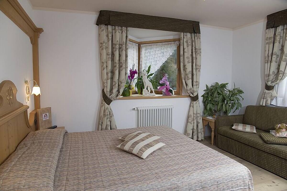 Un dormitorio con una cama grande, cabecera de madera, mesita de noche con lámpara, ventana con cortinas, radiador, sofá verde y planta en maceta.