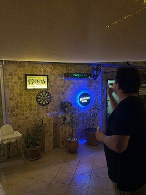 A man playing darts in a room with a dartboard, a sign reading "GUSTA", and a neon sign that says "#BIRKIE GÜZEL".