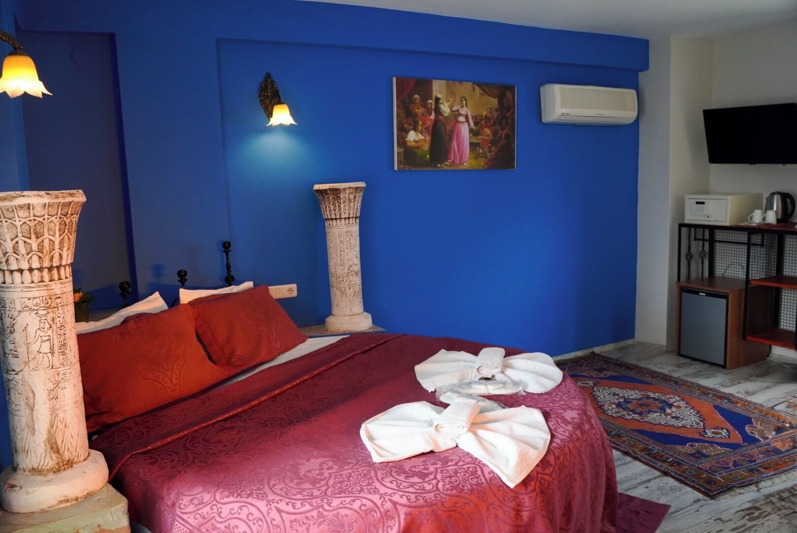 A room with a bed, a wall-mounted TV, a small cabinet, and a decorative pillar.