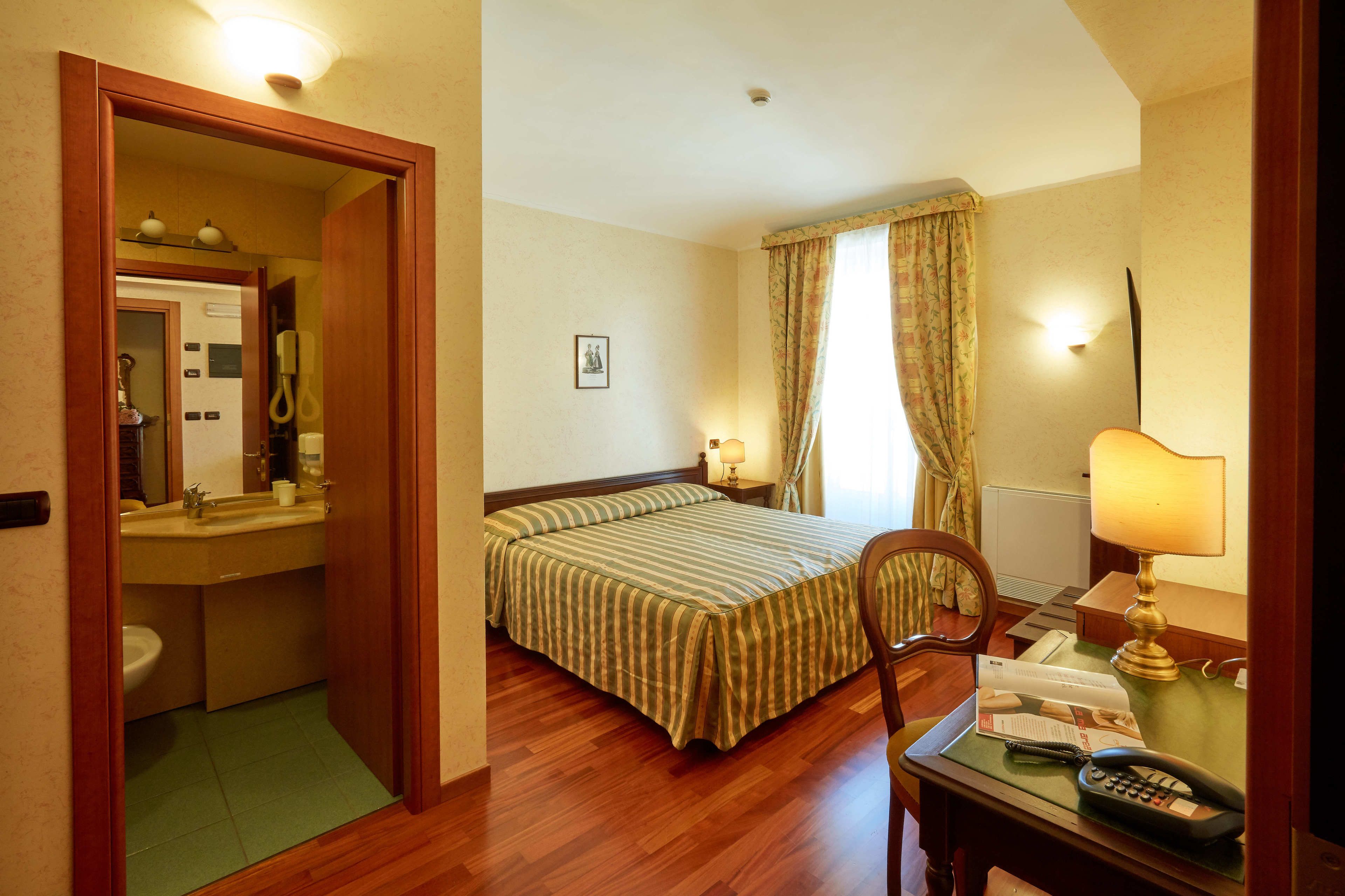 Une chambre d’hôtel avec un plancher en bois, un lit avec une literie à rayures, un bureau avec une lampe, un téléphone et une chaise.