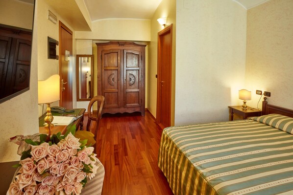 A hotel room with a wooden wardrobe, a bed with striped bedding, a desk with a lamp, and a bouquet of pink roses.