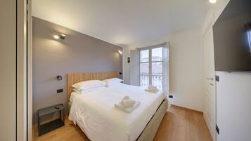 A modern hotel room with a large bed, a bedside table, a small desk, a window with a view, and a TV on the wall.