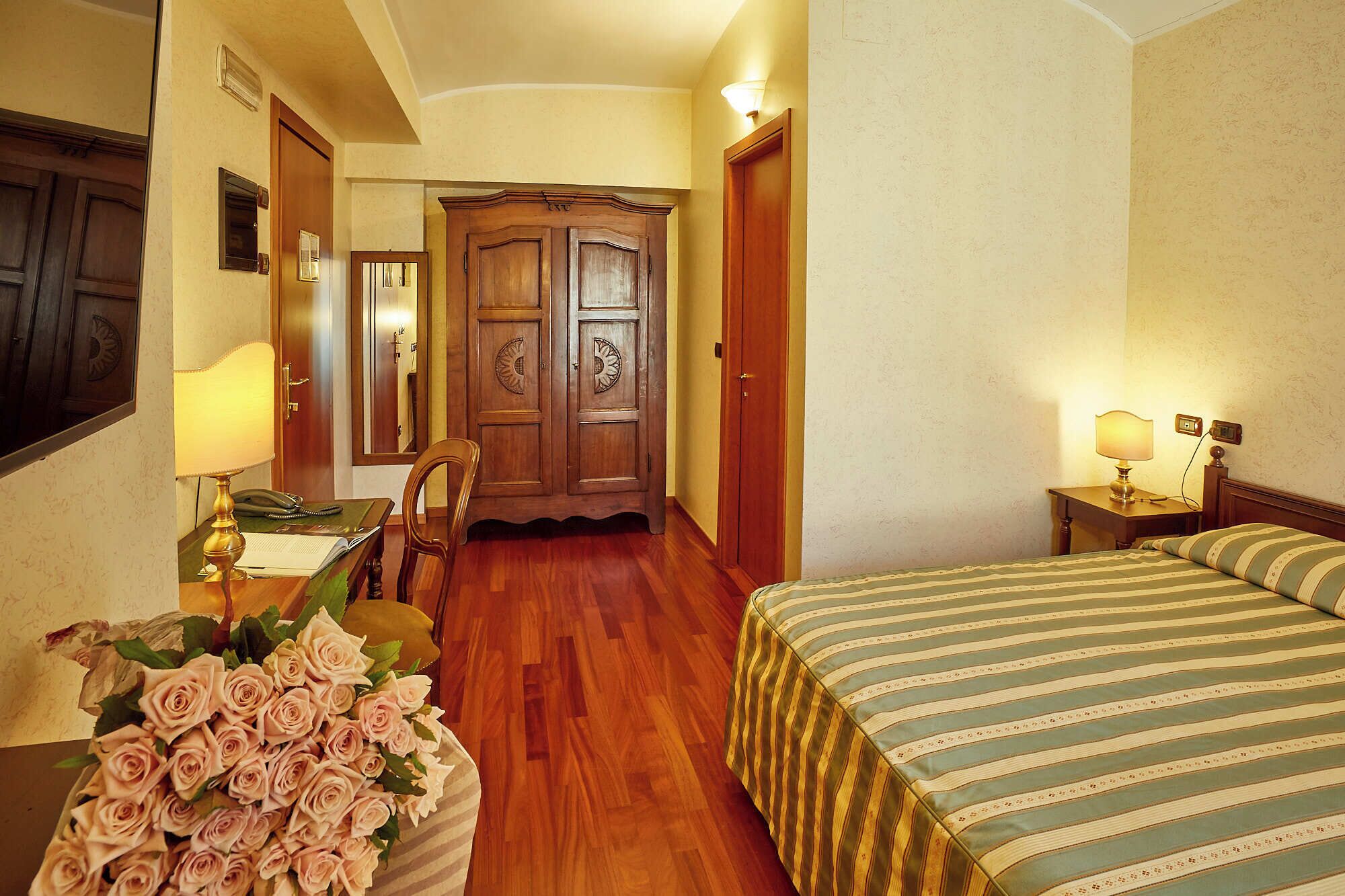 Une chambre d’hôtel avec une armoire en bois, un bureau avec une lampe, un lit avec une literie à rayures et un vase rempli de roses.
