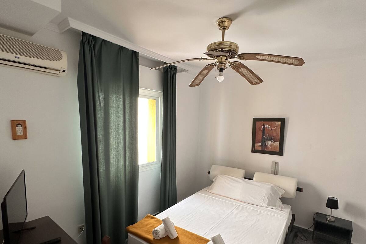 A hotel room with a bed, a desk, a chair, a ceiling fan, a picture on the wall, and a window with curtains.