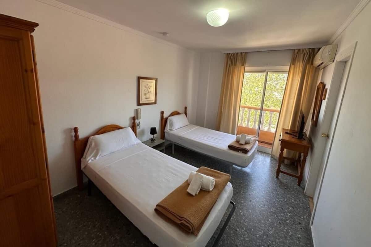 A hotel room with two single beds, a wooden wardrobe, a framed picture on the wall, and a window with curtains.