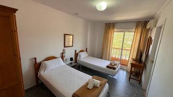 A hotel room with two single beds, a wooden wardrobe, a framed picture on the wall, and a window with curtains.