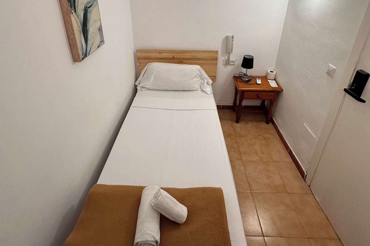 A single-bed room with a wooden headboard, a painting on the wall, a bedside table with a lamp, and a towel on the bed.
