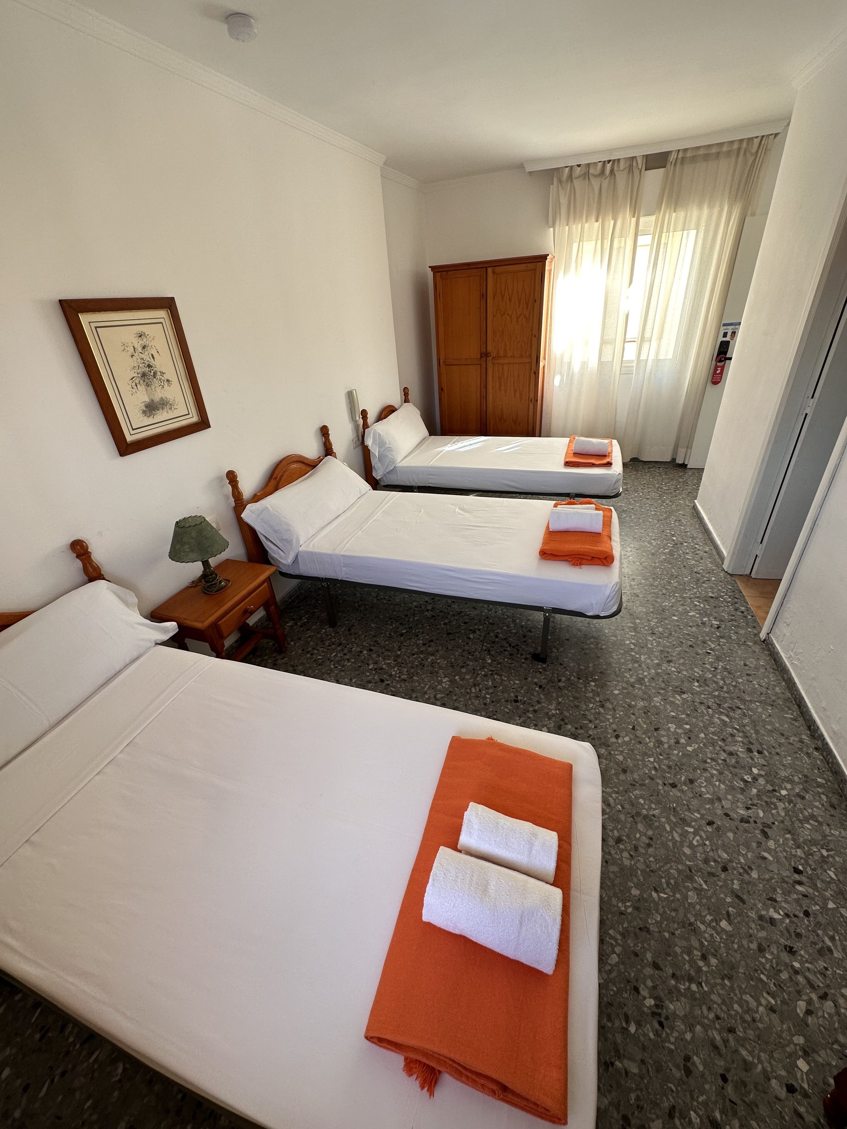 A hotel room with two beds, a wooden wardrobe, a framed picture on the wall, and a small table with a lamp.