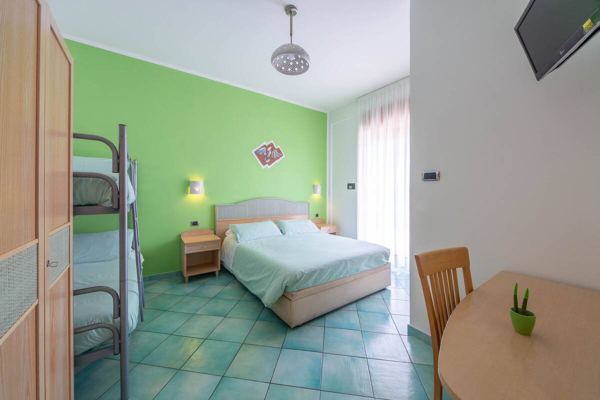 A bedroom with a bunk bed, a single bed, a bedside table with a lamp, a small table with a plant, and a TV mounted on the wall.
