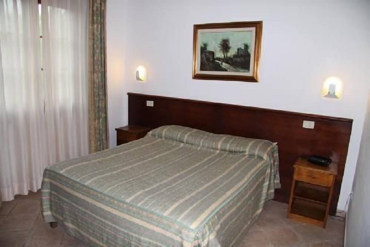 Ein Hotelzimmer mit einem Einzelbett, zwei Nachttischen, einem Fenster mit Vorhängen und einem gerahmten Bild an der Wand.