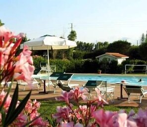 A pool area with lounge chairs and umbrellas, surrounded by greenery and a building in the background. - Hotel Tris (Orbetello)