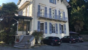 A two-story building with a balcony on the second floor, a staircase leading to the entrance, and a parking area with cars. - Hôtel Montilleul (Pau)
