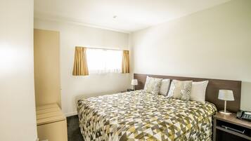 Quarto de hotel com uma cama grande, uma mesa de cabeceira com luminária, uma pequena escrivaninha com telefone e uma janela com cortinas.