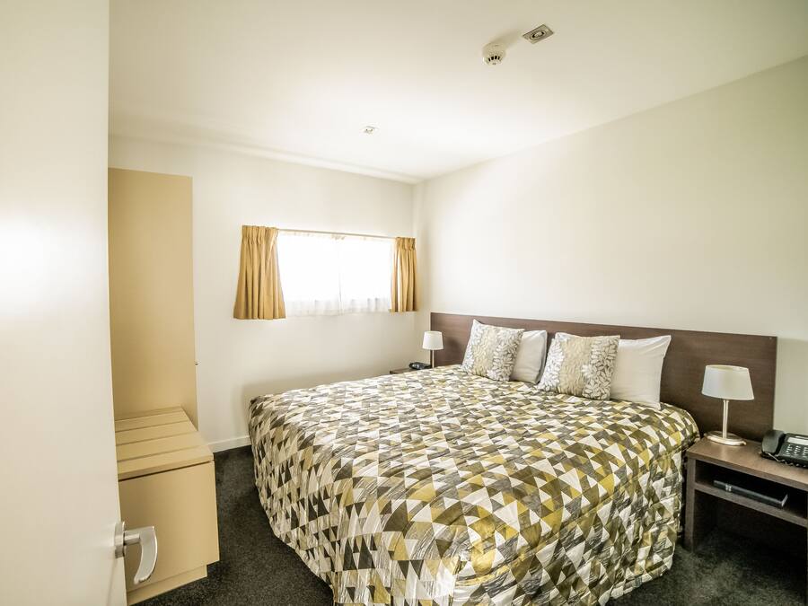 Quarto de hotel com uma cama grande, uma mesa de cabeceira com luminária, uma pequena escrivaninha com telefone e uma janela com cortinas.