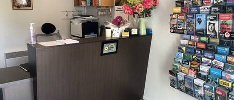 A reception desk with a clock, a small shelf with books and a vase of flowers, and a wall display of travel brochures.