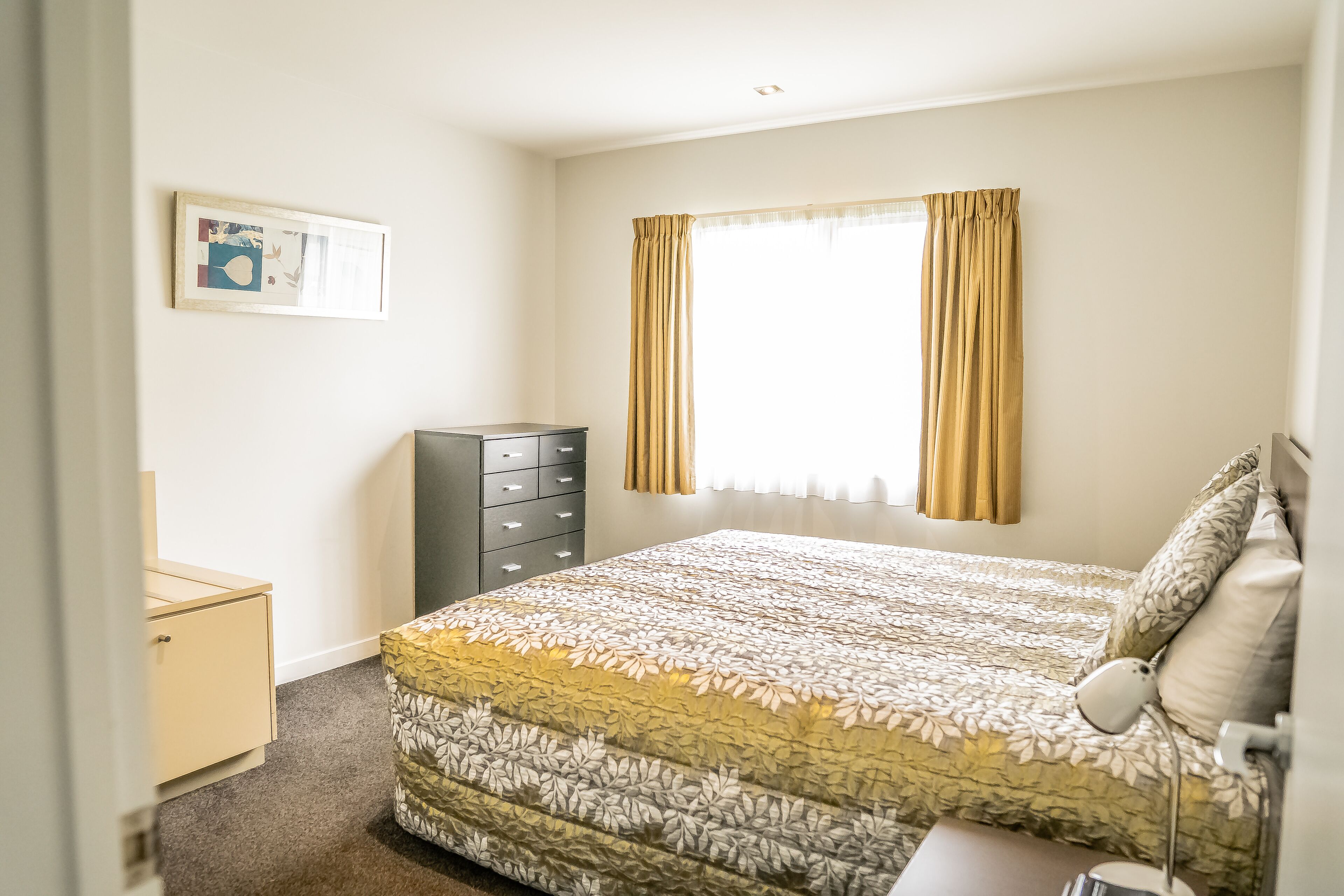 Quarto com cama, penteadeira, mesa de cabeceira, abajur e janela com cortinas.