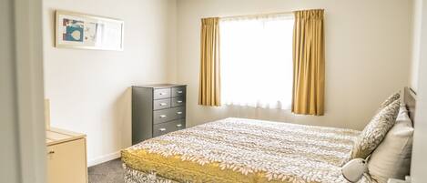 Une chambre à coucher comprenant un lit, une commode, une table de chevet, une lampe et une fenêtre avec des rideaux.