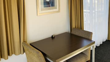 A hotel room with a wooden table, two chairs, a framed picture of tulips, beige curtains, and an air conditioning unit.