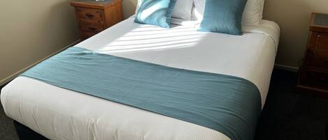 Una cama bien hecha con ropa de cama blanca y azul verdosa, una lámpara de noche y una mesita de madera.