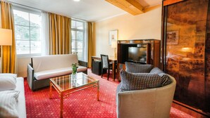 Habitación de hotel con alfombra roja, un sofá, una silla, un televisor y un ventanal con cortinas.
