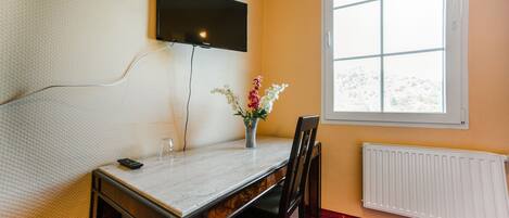 Una habitación con un televisor montado en la pared, un florero con flores sobre una mesa, una silla y una ventana que ofrece vistas de nieve.
