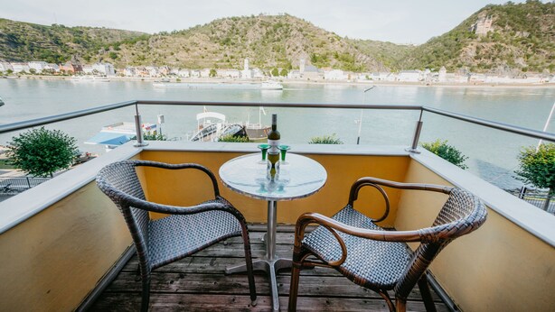 Un balcón con sillas de mimbre y una pequeña mesa, con vista a una masa de agua con barcos y un paisaje montañoso al fondo.