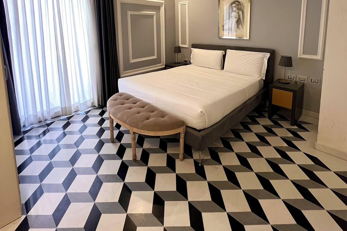 A hotel room with a large bed, a bedside table, a nightstand, a wall-mounted air conditioner, a window with curtains, and a geometric patterned floor.