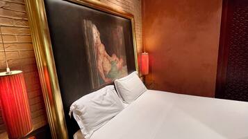 Habitación de hotel con pared de ladrillo, una cama grande con sábanas blancas, un cuadro enmarcado sobre la cama, una lámpara roja y un mueble de madera con cajones.