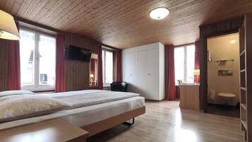 Une chambre d’hôtel avec un plafond en bois, un lit avec un matelas et des oreillers, un bureau avec une chaise, une armoire, un téléviseur fixé au mur et une salle de bain avec des toilettes et un lavabo.