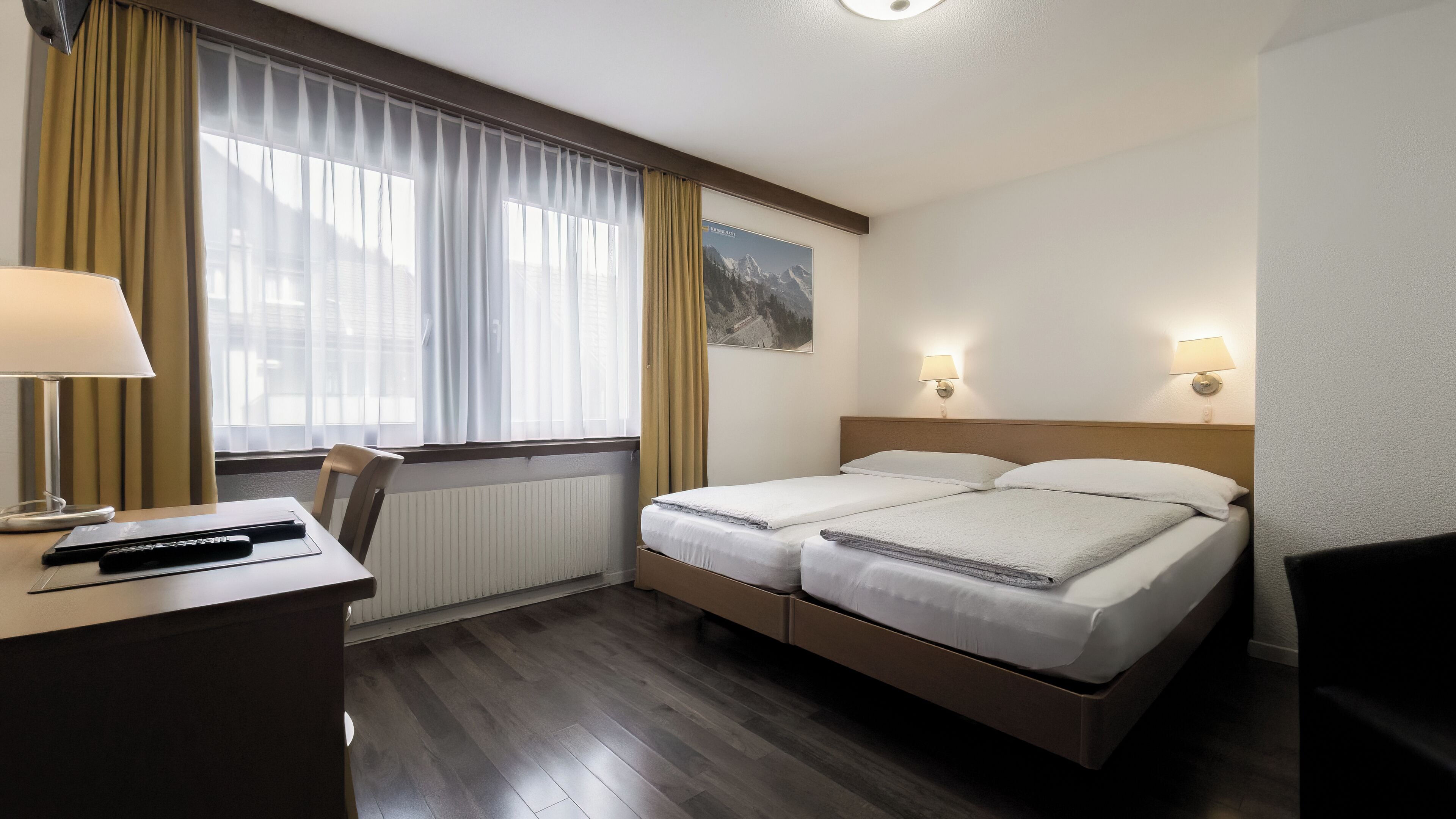 A hotel room with a double bed, a desk with a lamp, a chair, and a window with curtains.