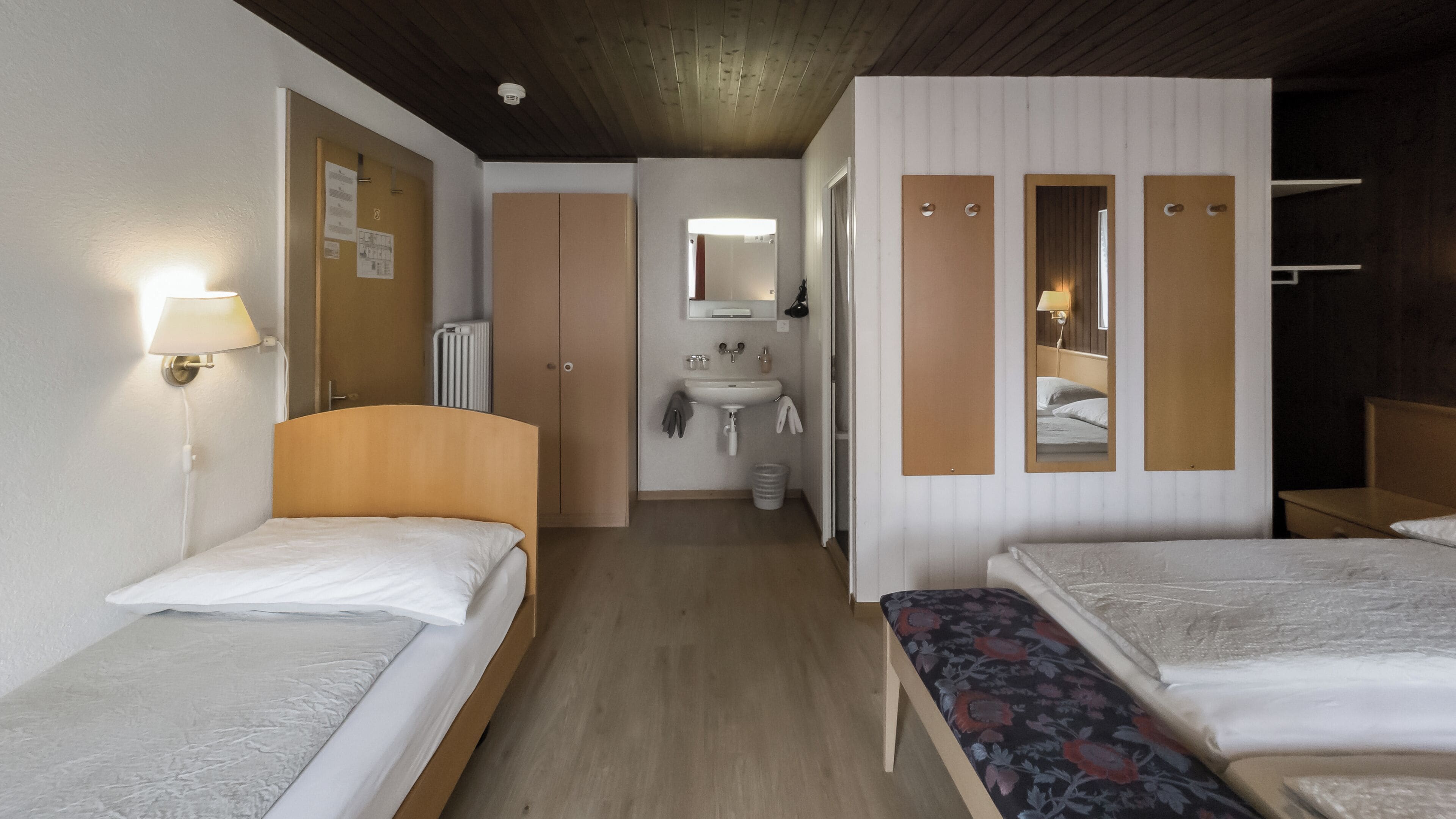 A hotel room with two single beds, a bathroom with a sink and mirror, and a wooden ceiling.