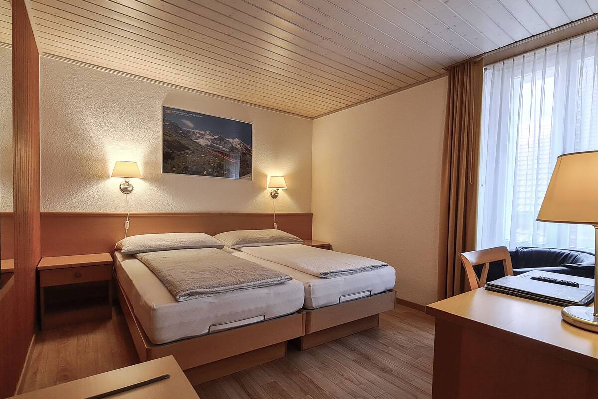 Habitación de hotel con dos camas, cabeceros de madera, un escritorio con una silla, una lámpara y un cuadro en la pared.