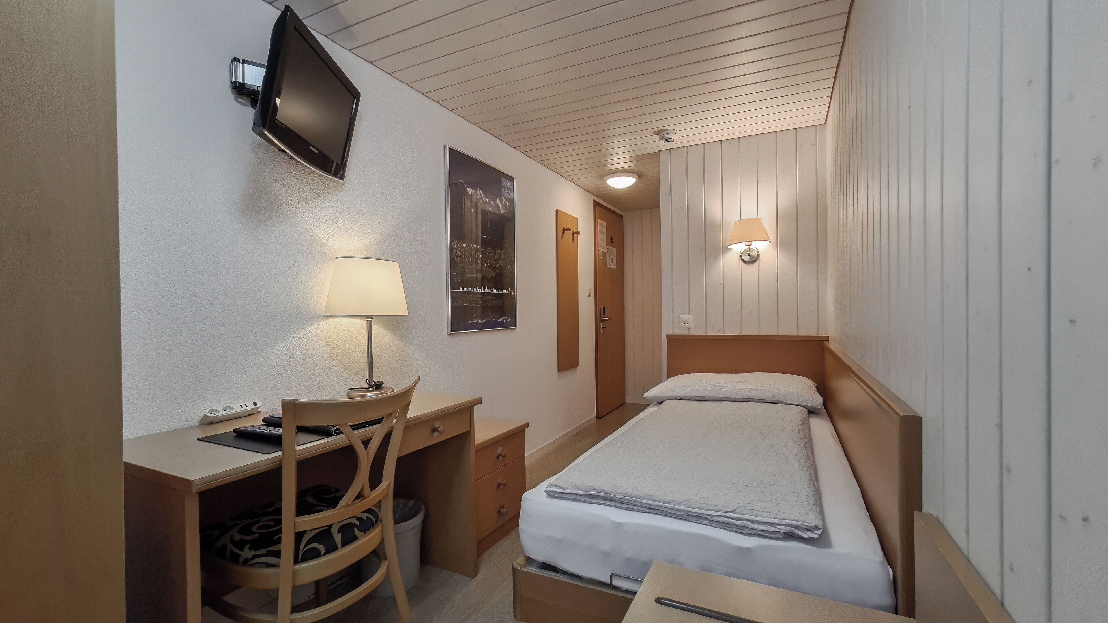 Una habitación compacta con cama, escritorio con silla, televisor montado en la pared y lámpara de pared.
