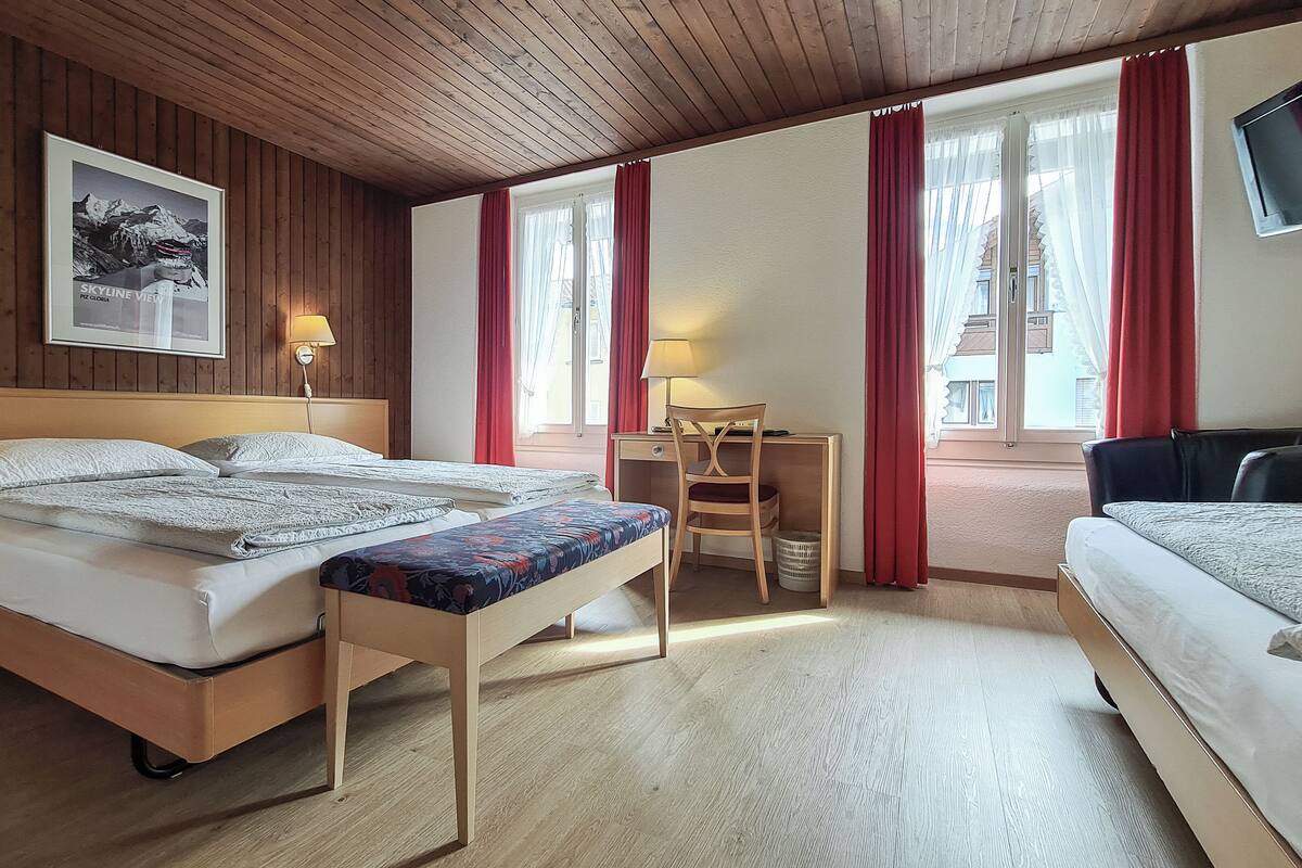 Habitación de hotel con techo de madera, cama con cubrecama blanco, escritorio con silla y televisor montado en la pared.