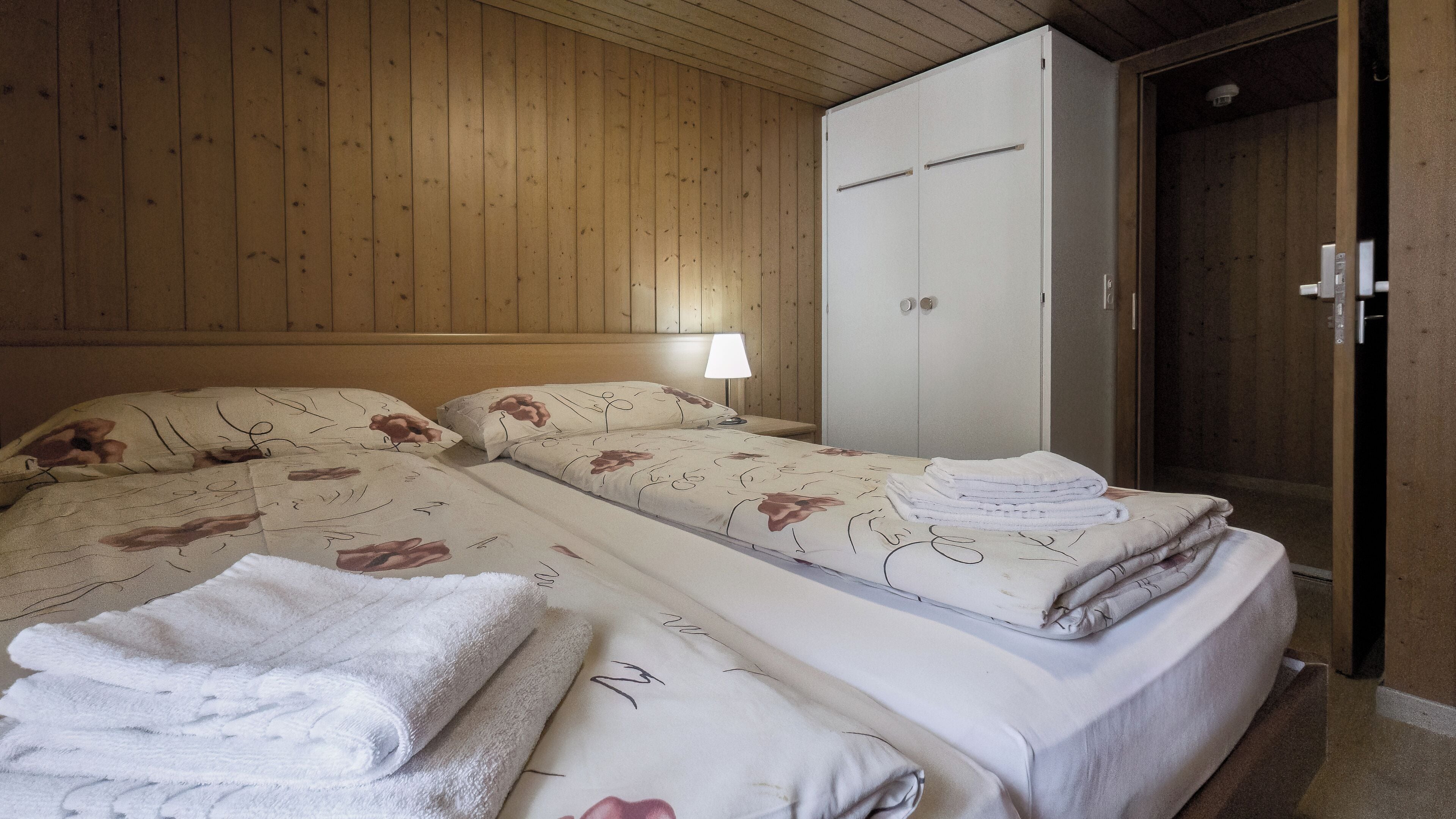 A bedroom with two beds, wooden walls, a wardrobe, and a nightstand with a lamp.
