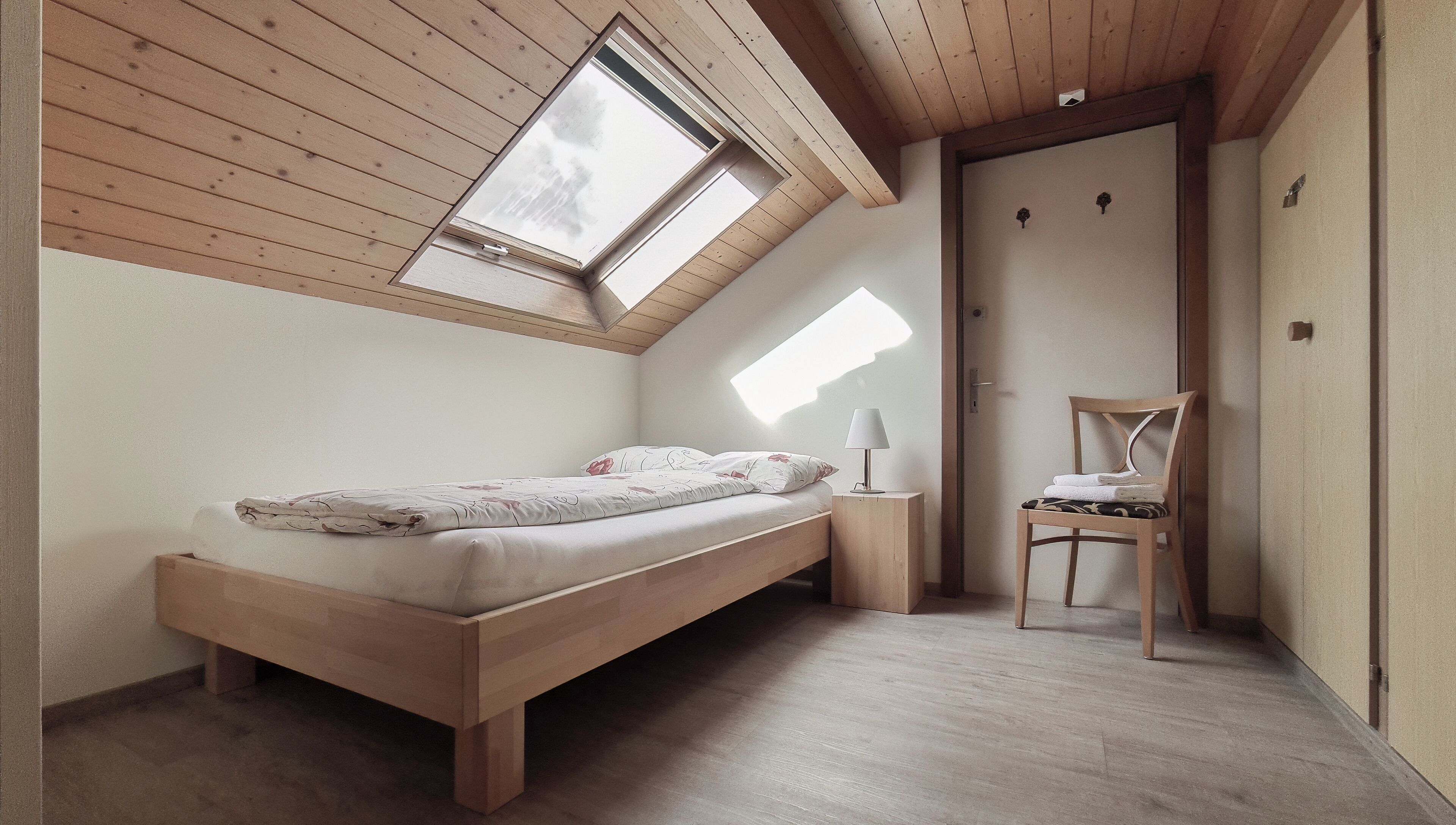 Un dormitorio con techo inclinado, vigas de madera, una cama, una silla y una mesita con lámpara.