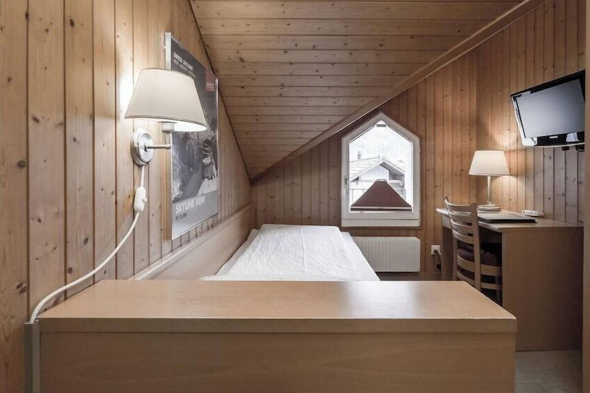 Una habitación pequeña y acogedora con paredes de madera, una cama individual, un escritorio con silla, un televisor montado en la pared y una ventana con vistas a un edificio.