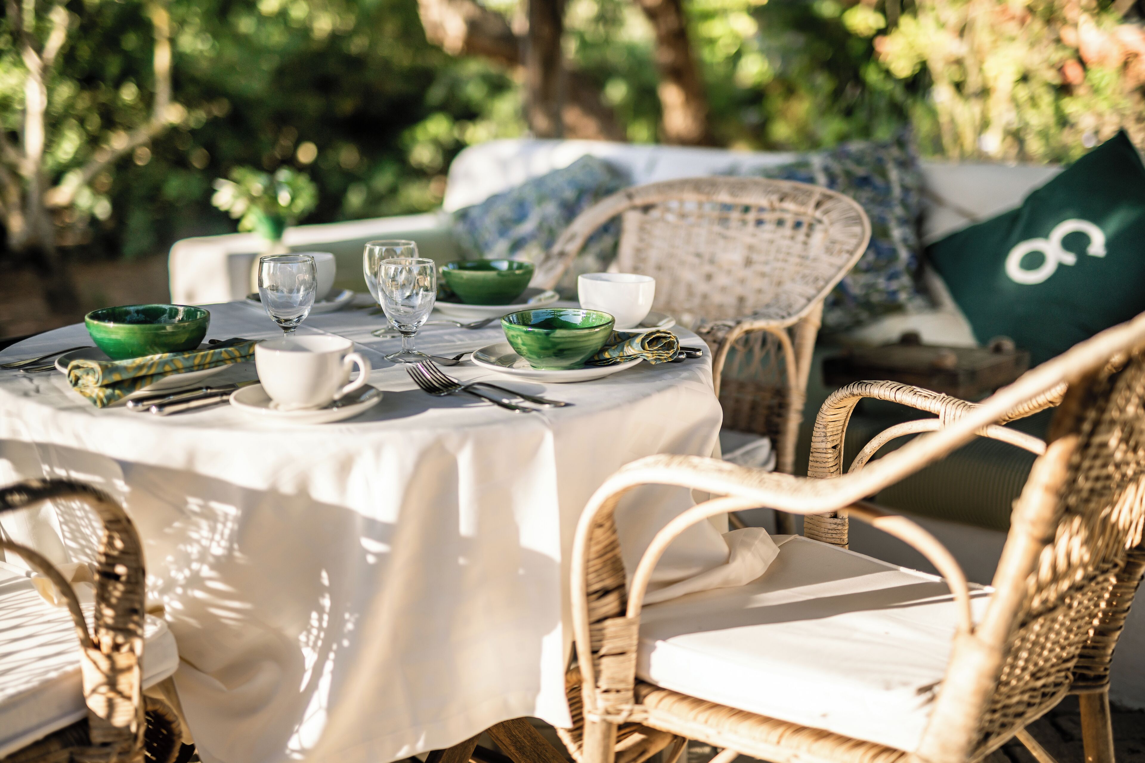 A round table set with green bowls, white cups, and cutlery, surrounded by wicker chairs and a white tablecloth.