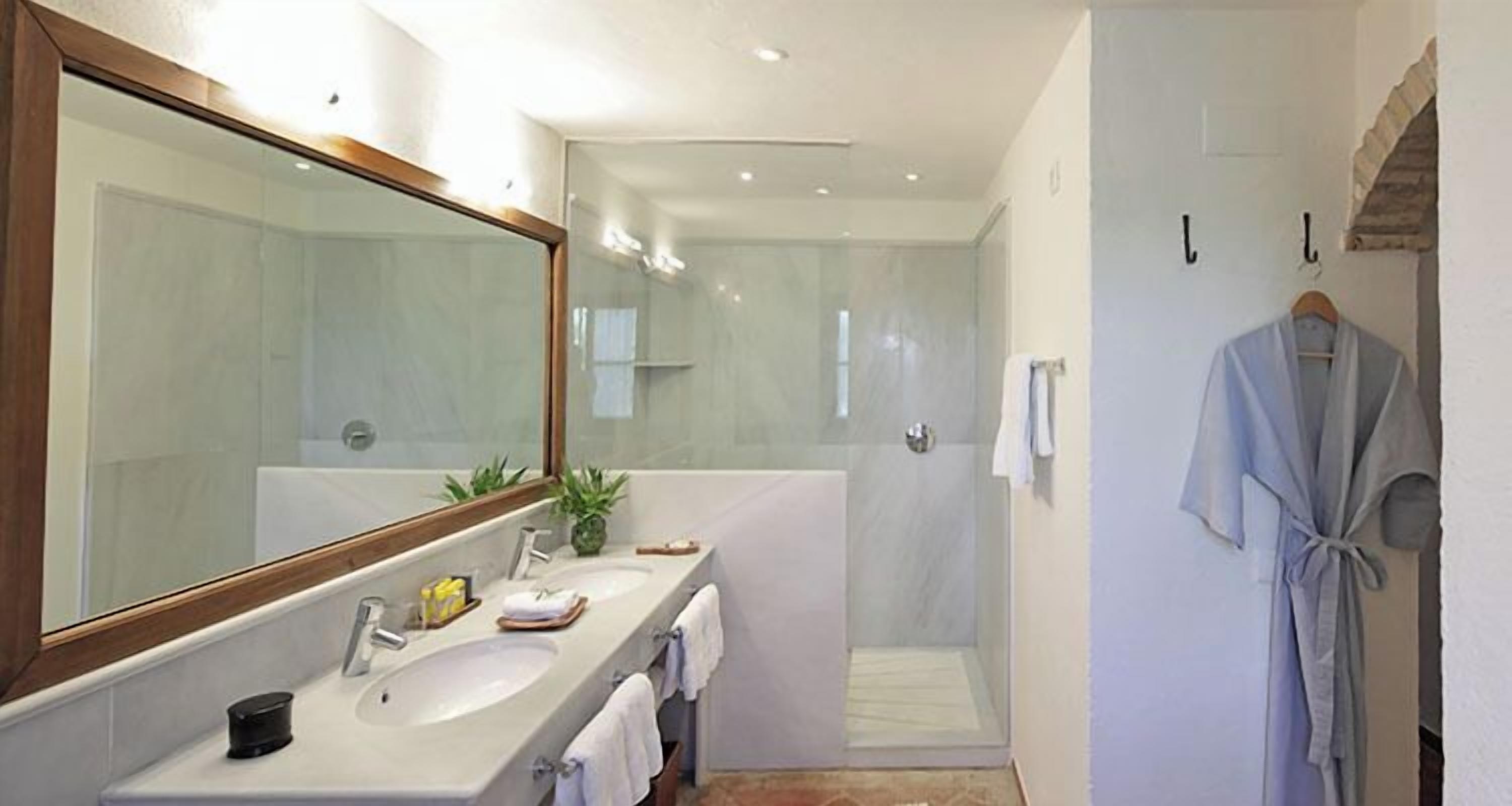 Un baño moderno con un espejo grande, dos lavamanos, una ducha y una bata colgada en la pared.