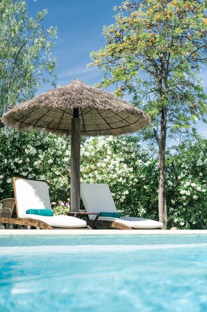 A pool area with two lounge chairs, a thatched umbrella, and lush greenery in the background.