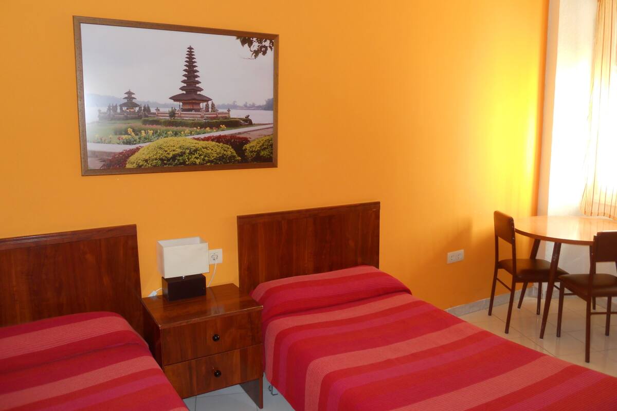A hotel room with two beds, a desk, a chair, and a wall-mounted picture of a pagoda.