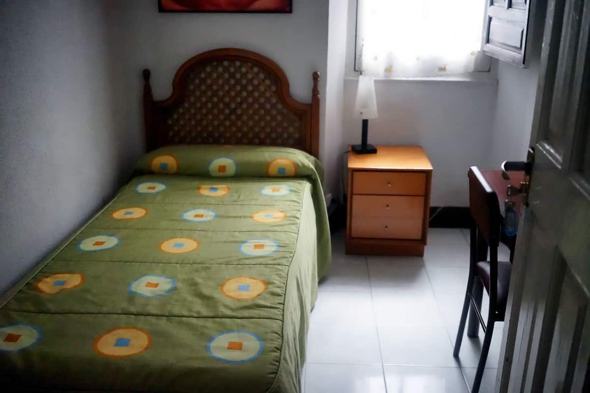 A small, neatly arranged bedroom with a single bed, a nightstand, a chair, and a window with curtains.