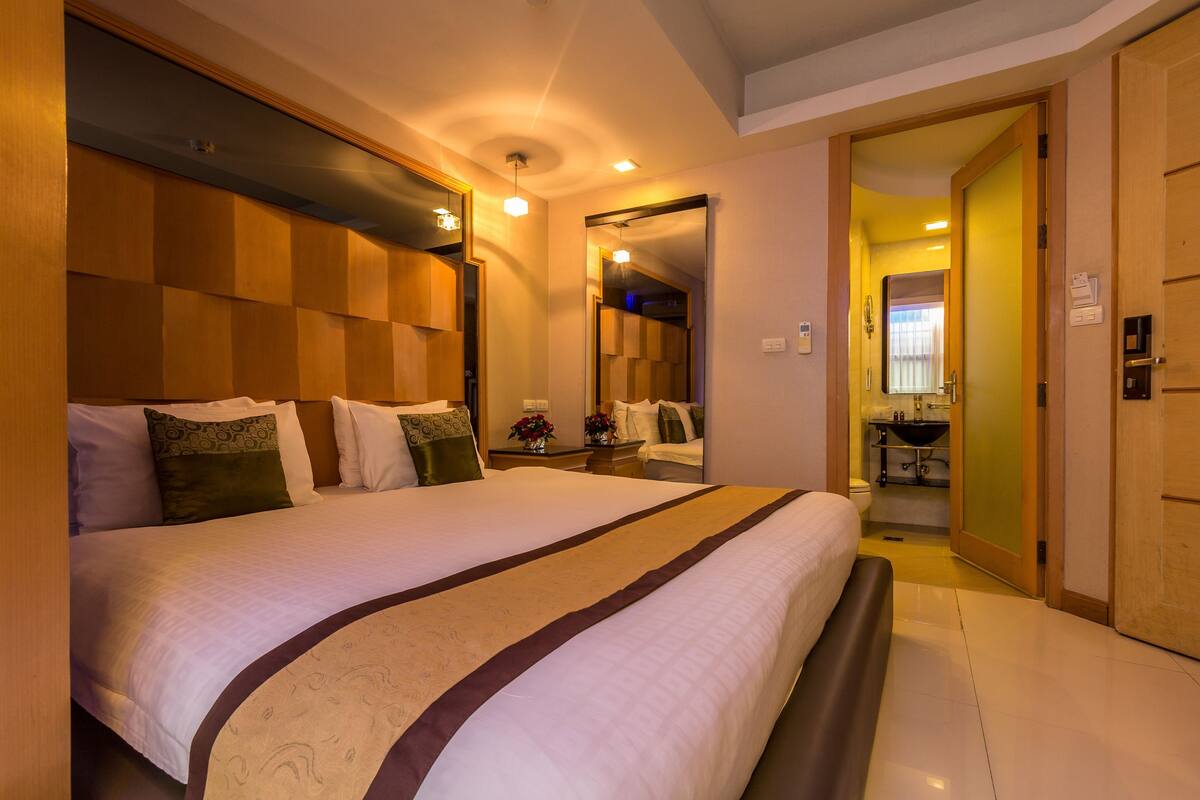 A hotel room with a large bed, a wooden headboard, a bedside table with a vase of flowers, and a bathroom visible through an open door.
