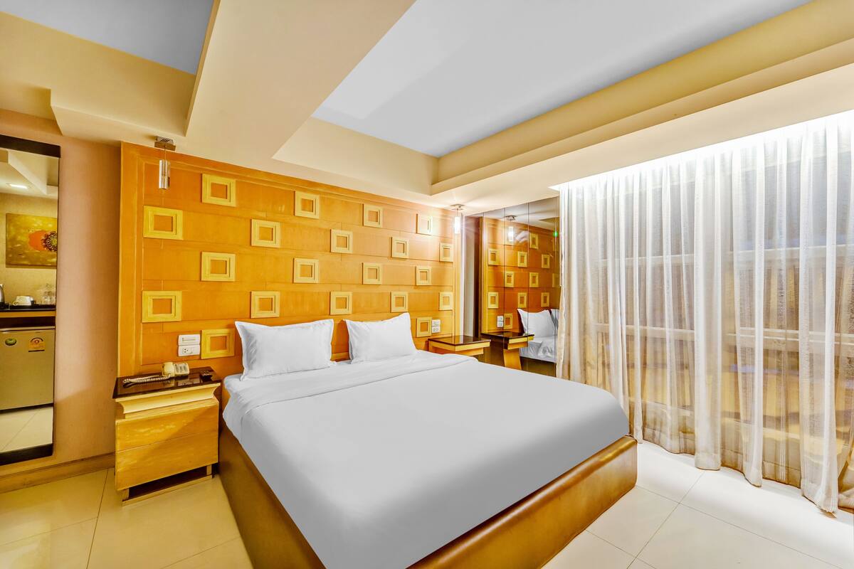 A hotel room with a large bed, wooden headboard, bedside tables, a mirror, and a window with sheer curtains.