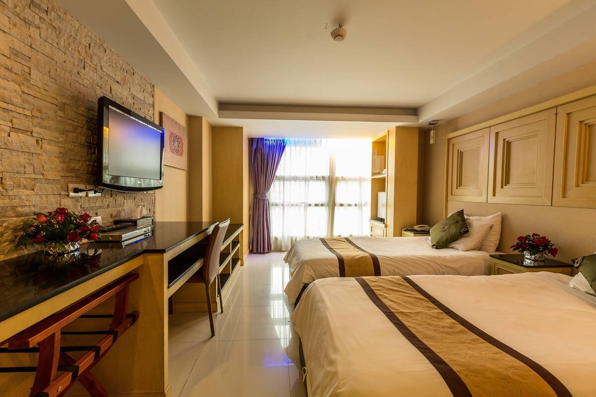 A hotel room with a brick wall, a flat-screen TV, a desk, two beds, a nightstand, and a window with curtains.